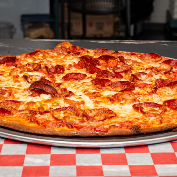A whole Halal Krush pepperoni pizza with melted cheese on a metal pan over a red and white checkered tablecloth.
