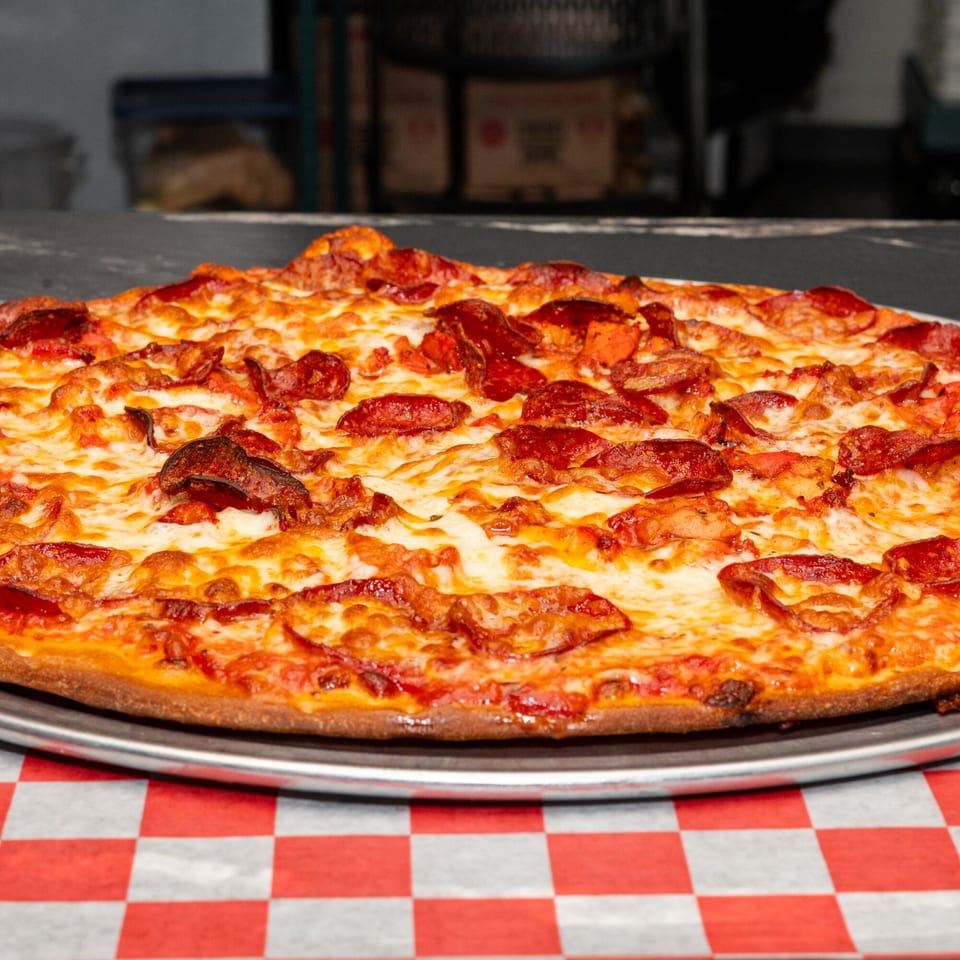 A whole Halal Krush pepperoni pizza with melted cheese on a metal pan over a red and white checkered tablecloth.